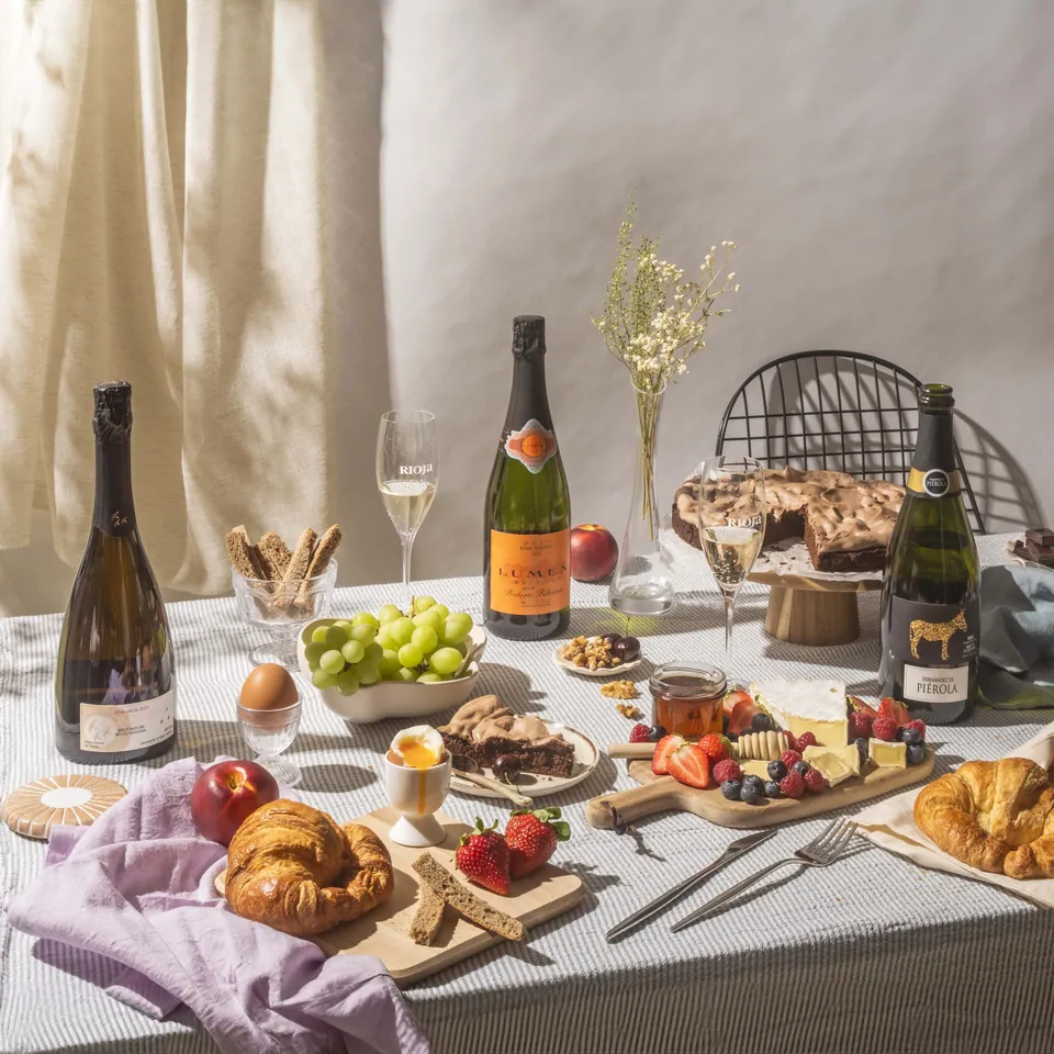 Mesa de picoteo con botellas de cava, frutas y cruasanes.