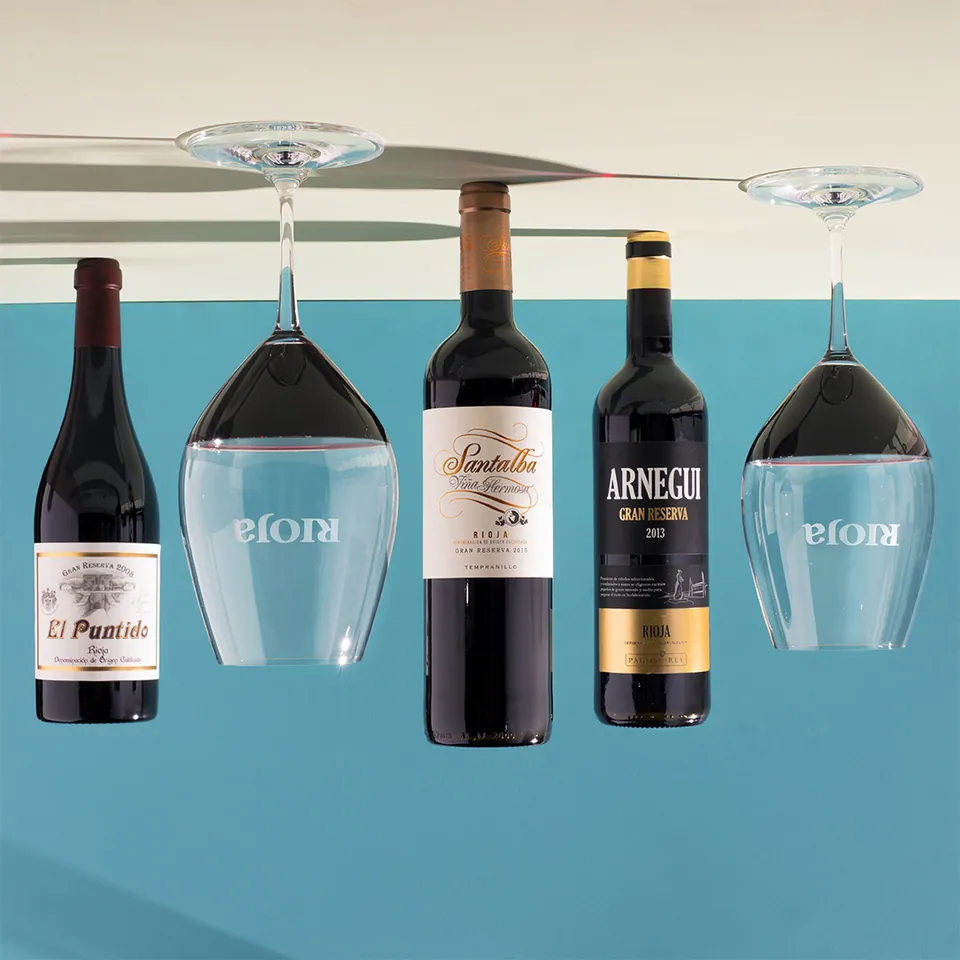 Botellas de vino tinto y copas colgadas boca abajo sobre fondo turquesa.