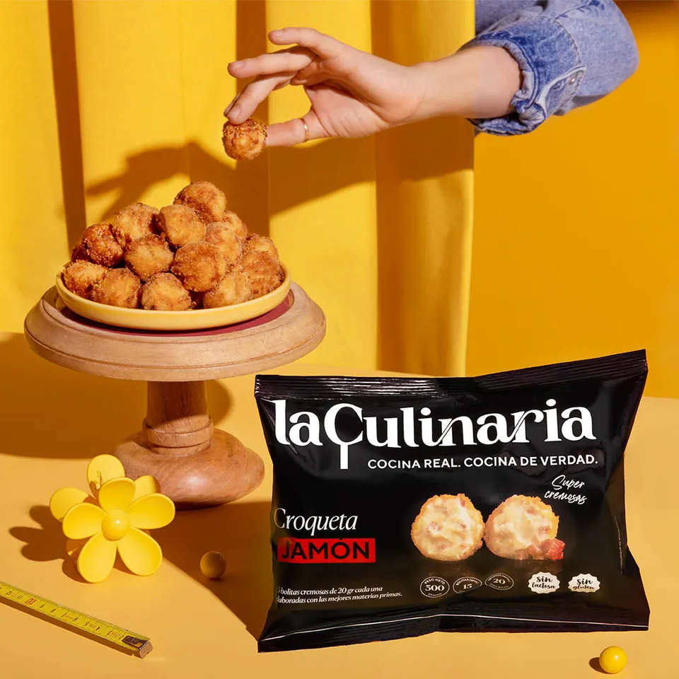 Mano que toma una croqueta sobre fondo amarillo, con la bolsa de La Culinaria.